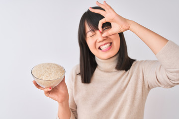 Young beautiful Chinese woman holding bowl with rice over isolated white background with happy face smiling doing ok sign with hand on eye looking through fingers