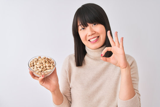 Young beautiful Chinese woman holding bowl with pistachios over isolated white background doing ok sign with fingers, excellent symbol