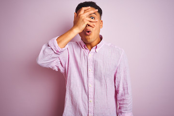 Young brazilian man wearing shirt standing over isolated pink background peeking in shock covering face and eyes with hand, looking through fingers with embarrassed expression.