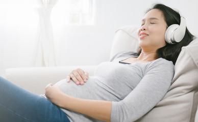 Portrait of pregnant asian woman sitting on sofa at home and listening music in headphones.