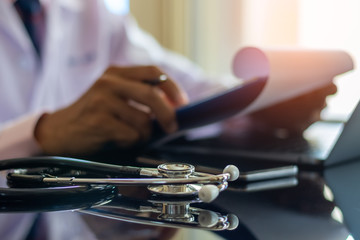 Male doctor work on laptop computer, hand holding clipboard with medical stethoscope on the desk at office.