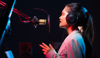 Asian Teenager Woman black hair earmuff headphone sing a song loudly power sound over hanging microphone condenser, speakers, composer note book and monitor. Studio Sound Proof Absorbing wall room