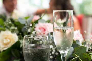 Copa de champán en primer plano con flores rosas y blancas y gente casual durante la fiesta y celebración de matrimonio, pareja hablando fuera de foco.
