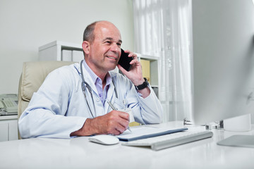 Smiling doctor reading information on computer screen and taking notes when talking on phone with patient
