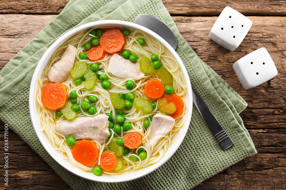 Wall mural Fresh homemade chicken noodle soup with carrot, peas and celery in white soup bowl, spoon on the side, photographed overhead