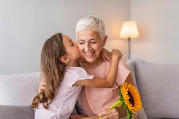 Foto op Canvas Happy senior grandma hugging granddaughter thanking for gift and flowers. Little granddaughter kissing giving flowers bouquet congratulating smiling old grandmother © Dragana Gordic