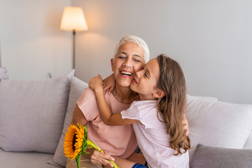  Happy senior grandma hugging granddaughter thanking for gift and flowers. Little granddaughter kissing giving flowers bouquet congratulating smiling old grandmother - obrazy, fototapety, plakaty
