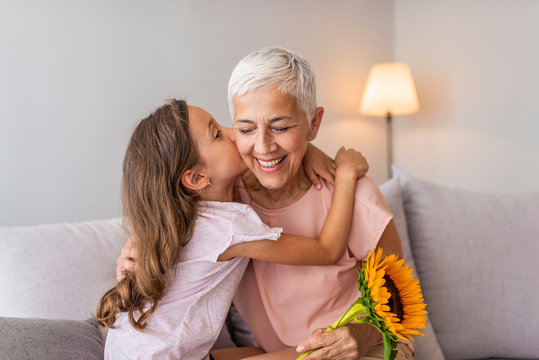 Happy senior grandma hugging granddaughter thanking for gift and flowers. Little granddaughter kissing giving flowers bouquet congratulating smiling old grandmother