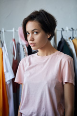 Beautiful young woman with short black hair wearing pink T-shirt standing in front of rack with hangers. close up portrait.sleepy girl standing near her wordrobe