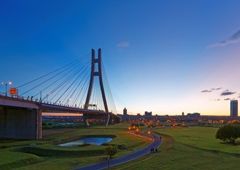Scenery of the riverside park by Tamsui River with a pathway thru the green grassy meadow & the grand bridge tower standing tall under beautiful sunset sky in New Taipei City, Taiwan (Low Angle View )