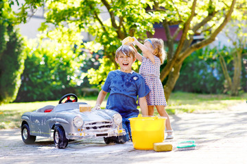 Two happy children washing big old toy car in summer garden, outdoors. Brother boy and little sister toddler girl cleaning car with soap and water, having fun with splashing and playing with sponge.