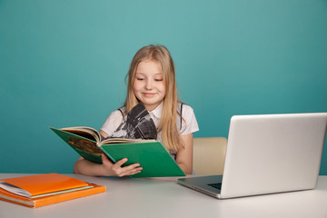 Little girl sitting at the desk and reading book infront of the laptop