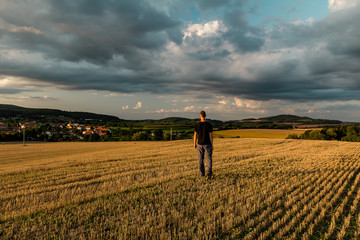 Young man walking on mowed field with dramatic sky at sunset, Czech republic