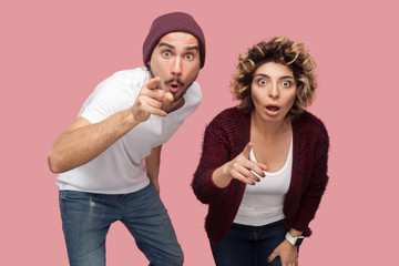 What? Portrait of shocked couple of friends in casual style standing and pointing finger to you with big eyes and opened mouth, looking at camera. Isolated, indoor, studio shot, pink background