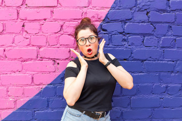 Portrait of a crazy young positive student girl in stylish clothes and glasses posing on a blue-violet brick background