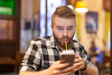 Shocked young hipster man drinking cocoa from a straw and looking at messages in social networks while sitting at a table in a cafe. The concept of chatting and social networks.
