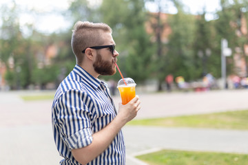 Portrait of a positive cheerful young man with a glass of juice with a straw while walking in the park on a warm sunny summer day. The concept of rest after study and work on weekends.