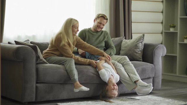 Playful family of three having fun together sitting on sofa in living room. Mother and father tickling little daughter laughing joyfully
