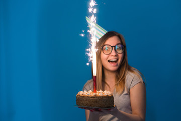 Cheerful young blurred girl student in glasses holding a congratulatory cake with a candle standing on a blue background. Birthday concept.