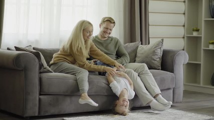Tracking left shot of family of three spending leisure time together at home. Mother and father sitting on sofa and tickling playful little daughter laughing cheerfully