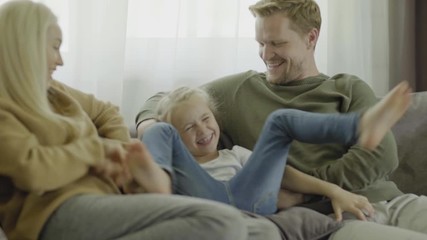 Handheld shaky camera shot of family of three having fun together at home. Mother and father sitting on sofa and tickling playful little daughter laughing joyfully
