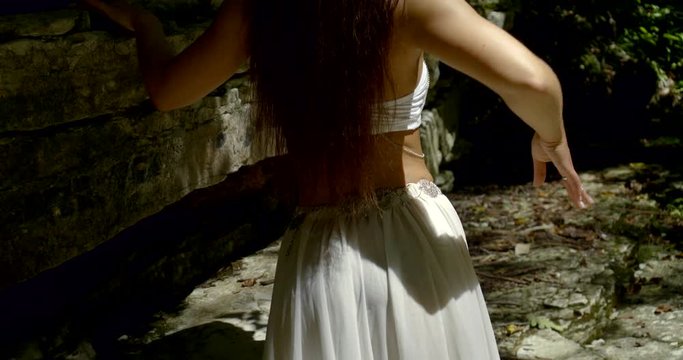 sexy young woman is wearing long white skirt is walking in forest with rocks and waving her hips