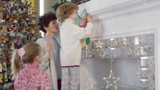 Tilt up of little boy standing on wooden step ladder and putting handmade paper stocking on fireplace with help of mother and sister while decorating home for Christmas