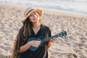 Woman with long hair in hat playing ukulele at the beach.
