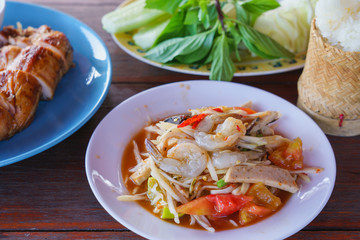Spicy papaya salad ( Som tum ) raw shrimp with white pork sausage on dish, Half grilled chicken, Sticky rice in bamboo container, vegetable on wooden table. Traditional Thai Asian food.