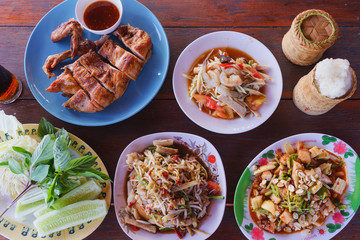 Spicy papaya salad ( Som tum ) raw shrimp with white pork sausage on dish, Half grilled chicken, Sticky rice in bamboo container, vegetable on wooden table. Traditional Thai Asian food. Top view