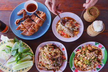 Spicy papaya salad ( Som tum ) raw shrimp with white pork sausage on dish, Half grilled chicken, Sticky rice in bamboo container, vegetable on wooden table. Traditional Thai Asian food. Top view