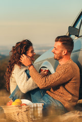 Beautiful young couple enjoying picnic time on the sunset. They drinking tea and sitting in a meadow leaning on old fashioned car.