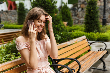 Portrait of excited laughing woman talking on cellphone while sitting on bench in summer park