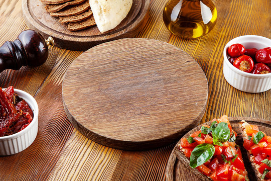 Top view empty cutting board for pizza or meat. Blank board for serving food in composition with, sun-dried tomatoes, bruschetta and utensils on wooden background. Flat lay
