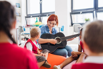 A group of small school kids and teacher with guitar sitting on the floor in class.