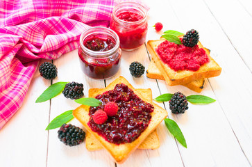 Toasters with blackberry and raspberry jam. Decorated with fresh berries and leaves of blackberry and raspberry on a white wooden table.