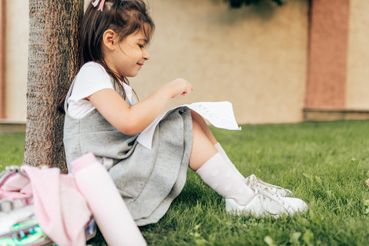 Side view image of cute little girl smiling and learning something on the paper, sitting under the tree on green grass, with backpack outdoors. Child pupil, relaxing after school day next to her house