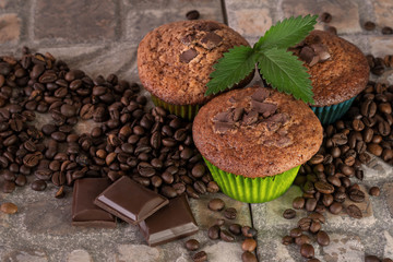 Three muffins with a green sprig and pieces of chocolate surrounded by coffee beans on an earthenware ground.