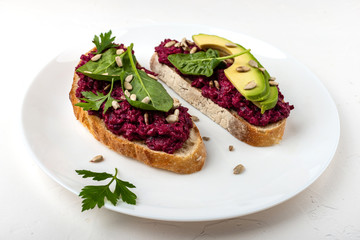 Bruschettas with beetroot hummus, avocado, spinach and seeds on a white plate on a white background.