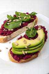 Bruschettas with beetroot hummus, avocado, spinach and seeds on a white plate on a white background.