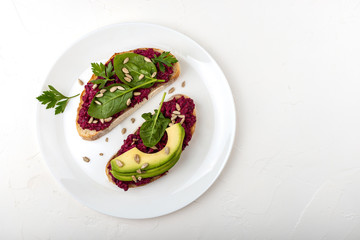 Bruschettas with beetroot hummus, avocado, spinach and seeds on a white plate on a white background.