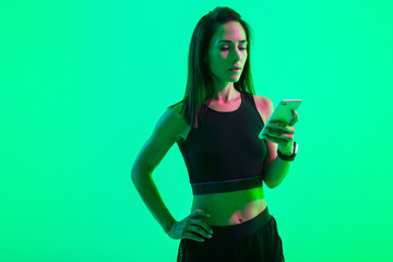 Young concentrated strong sports woman posing isolated over blue wall background with neon bright lights using mobile phone chatting.