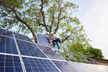 Two technicians mounting heavy solar photo voltaic panel on tall steel platform on green tree and...