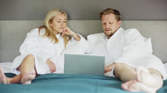 Beautiful middle aged couple in white bathrobes lying on bed in hotel and talking. Man using Internet on laptop computer