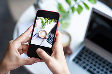 Mobile female taking photo of cappuccino in mug and laptop keypad on table