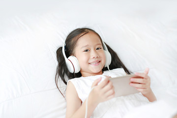 Happy little Asian girl using headphones listen music by smartphone smiling and looking camera while lying on bed at home.