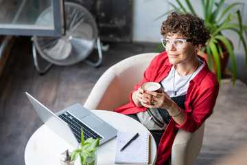 Young female designer with cup of coffee sitting in armchair in cafe