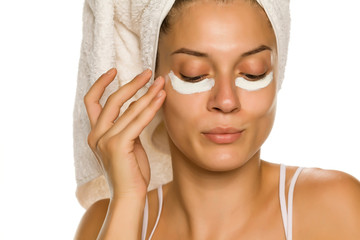 Young woman applying face cream under her eyes on white background