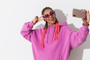 Smiling young african woman isolated over white wall background in bright pink sweatshirt wearing sunglasses take a selfie by mobile phone.