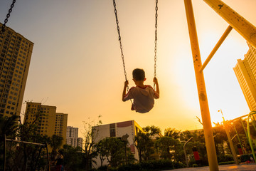 Child swinging on swing in sunset in city with building on background - obrazy, fototapety, plakaty